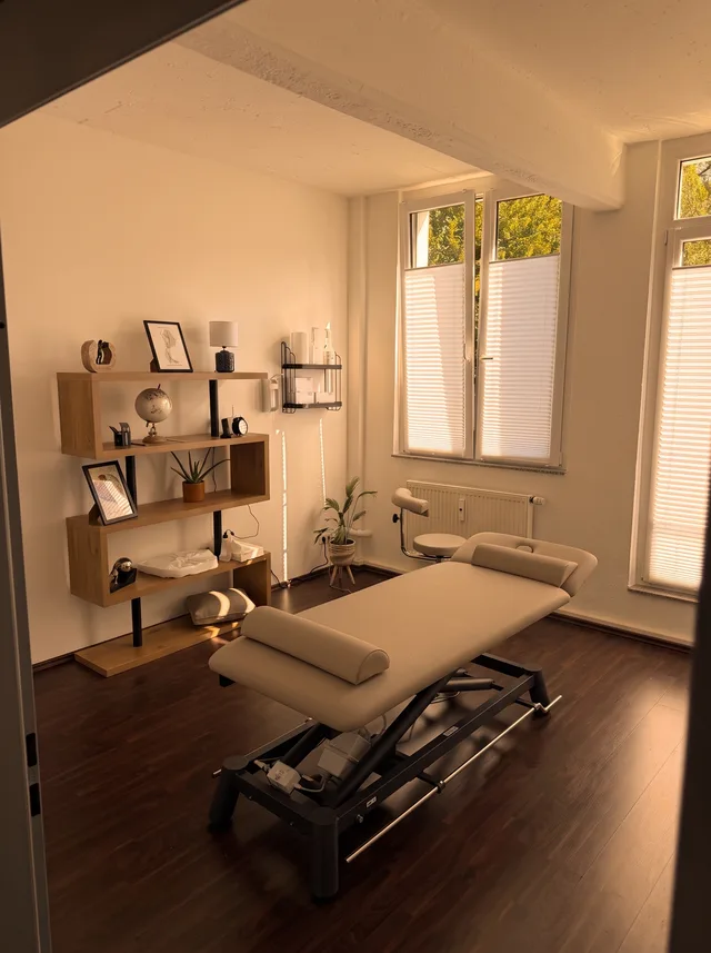Behandlungsraum mit Liege, offenem Wandregal und Fenster unter den Deckenbalken