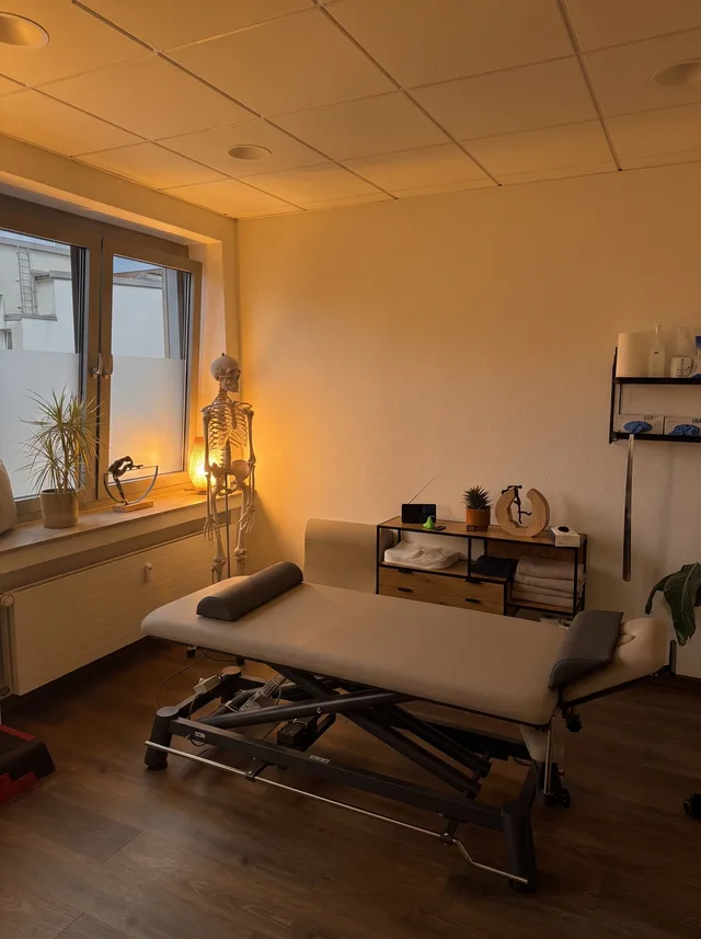 Behandlungsraum mit Liege, Skelettmodell am Fenster und einem Regal mit Handtüchern
